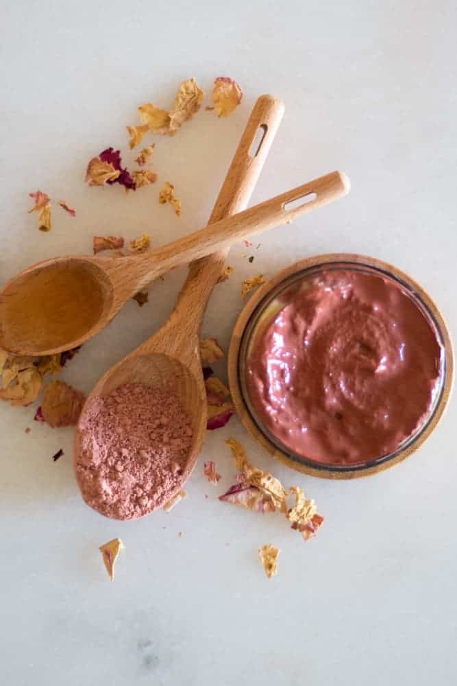 homemade rose clay face mask on white marble with rose petals and wooden spoon of rose clay 