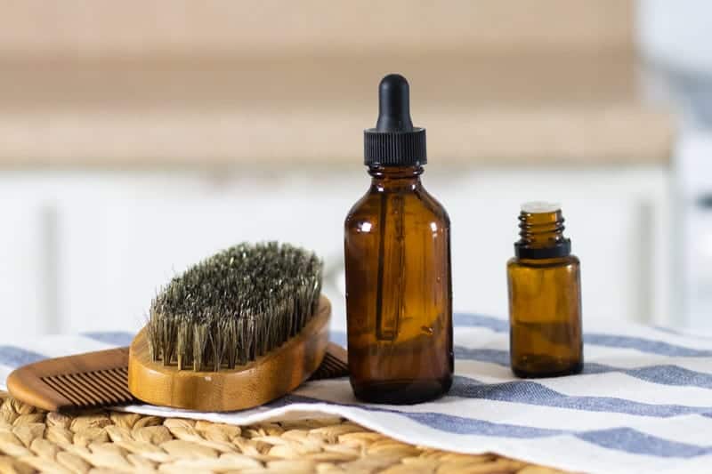 beard shampoo, amber colored essential oil bottle, and beard brush and comb on white and blue tea towel