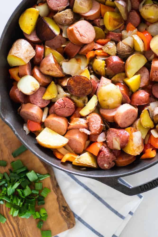 Crispy potatoes with chopped sausage links peppers and onions in a lodge skillet with a wooden cutting board of diced green onions.