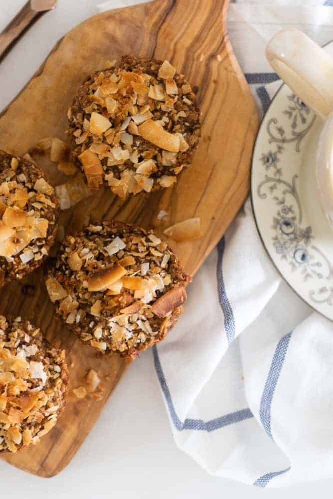 toasted coconut and applesauce muffins on wooden cutting board on white and blue striped towel next to a cup of coffee