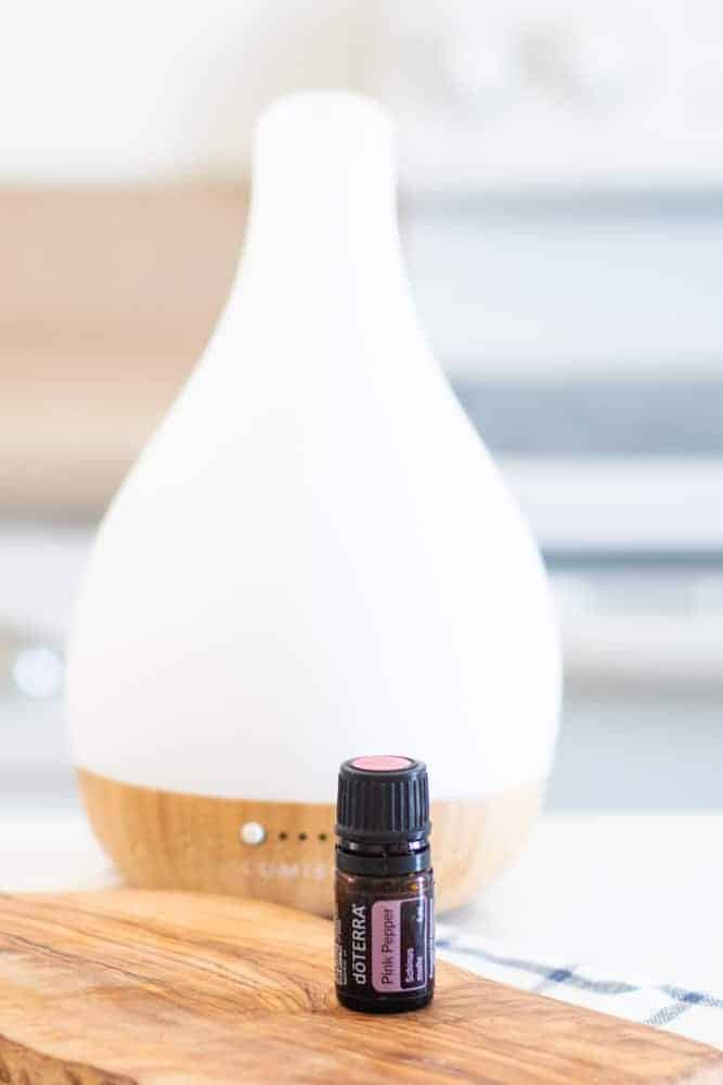 Doterra pink pepper bottle on wooden cutting board with a diffuser behind it. 