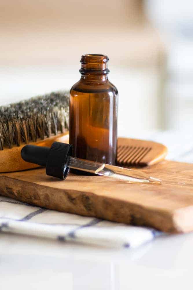 Dropper bottle with essential oils in it on wood board with bread brush and comb behind it. 