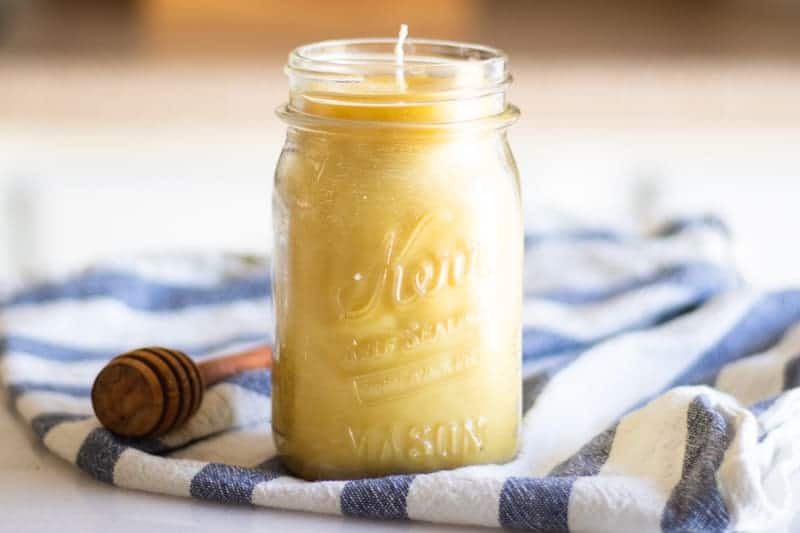 Homemade honey vanilla beeswax candle on table with white and blue tea towel. 