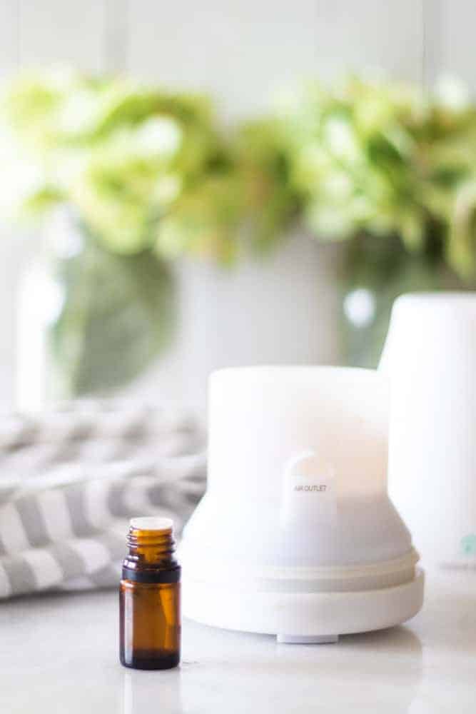 White diffuser on table with green flowers behind it. 