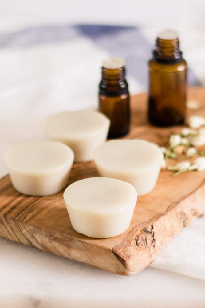 Homemade lotion bars on wood cutting board with amber colored essential oil bottles behind them. 