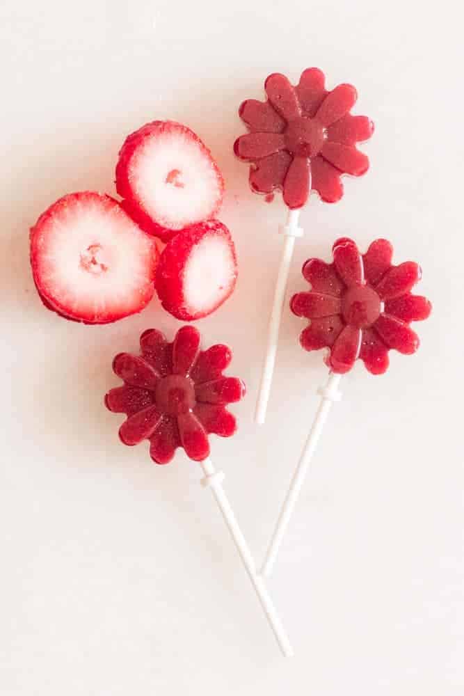 Homemade strawberry lollipops on white table with strawberry slices next to them.