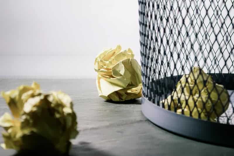 Metal trashcan with paper crumbled up next to it. 