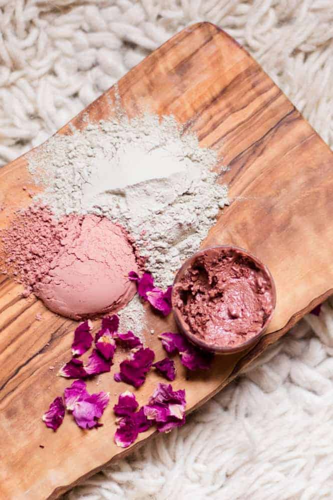 Rose clay, bentonite clay, and rose petals on wooden board with a homemade rose clay face mask in small glass container.