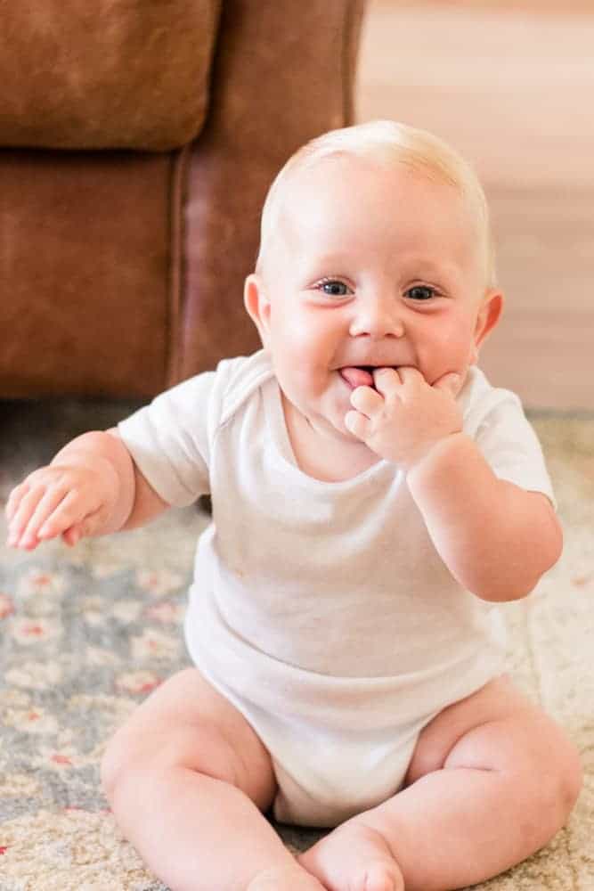 7 month old baby in white onesie sitting up sucking on fingers. 