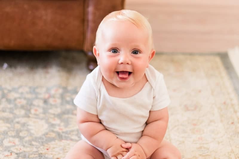 7 month old baby wearing a white onesie sitting on rug.