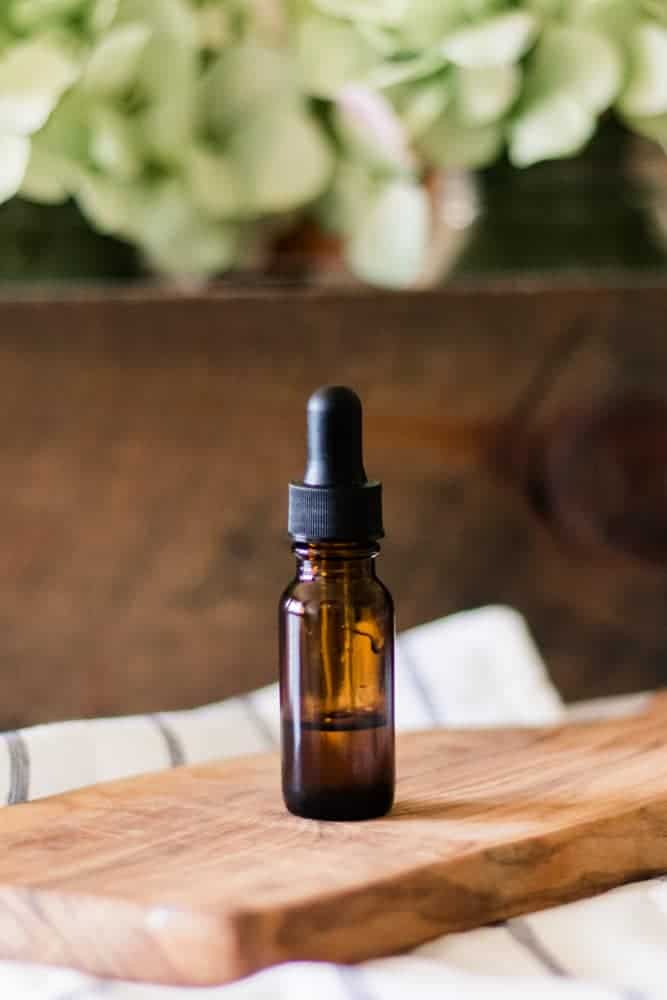 Vitamin E oil bottle standing on wooden cutting board with green flowers behind it. 