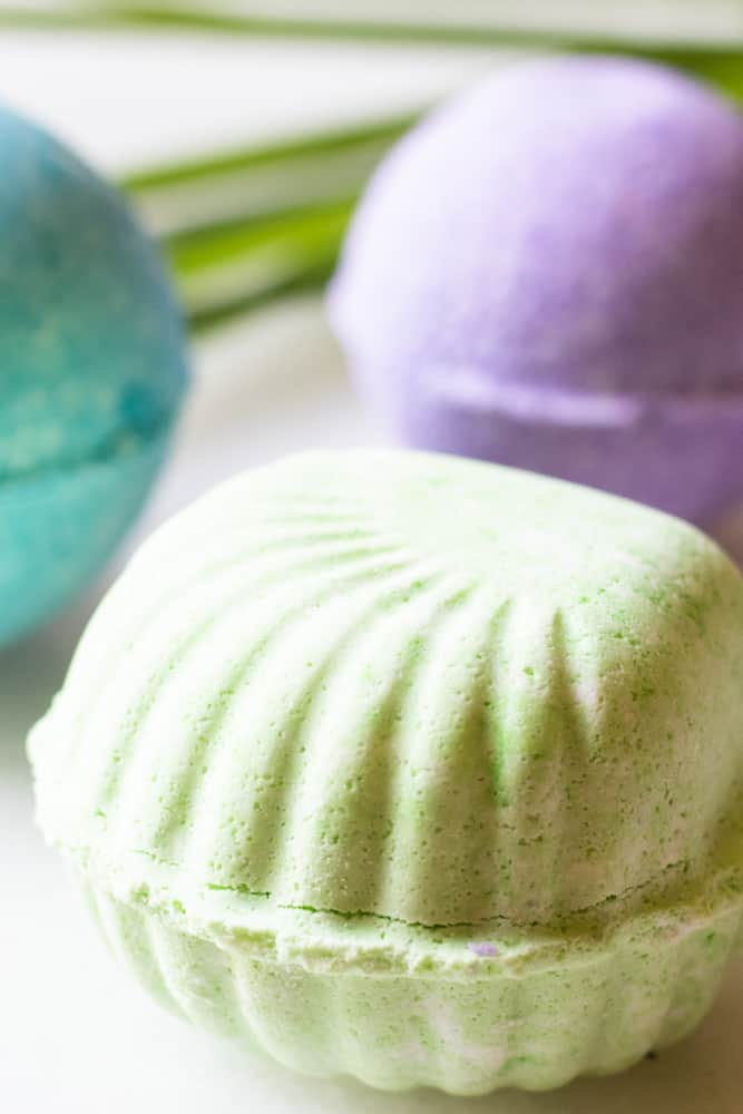 Seashell shaped green bath bomb with purple and blue bath bombs behind it.