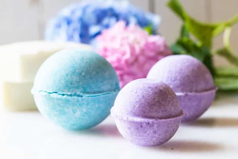 Bright purple and blue bath bombs on white table with flowers and soap bars behind them. 