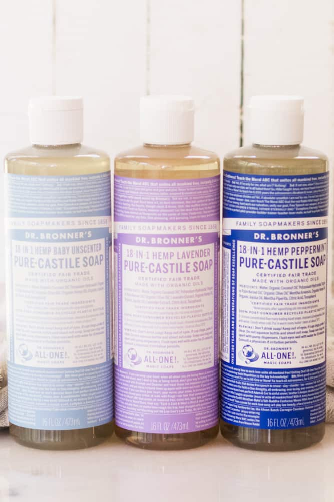 Three bottles of Dr. Bronners Castile Soap standing on white marble with white shiplap background.