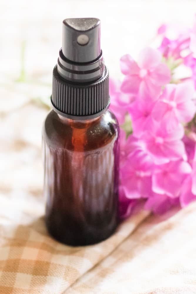 Amber glass spray bottle with homemade bathroom air freshener in it, fresh pink flowers sitting behind it.