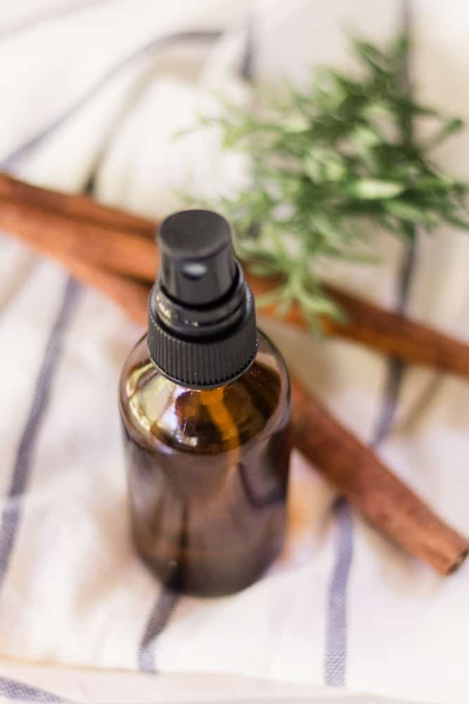 Glass spray bottle with cinnamon sticks and rosemary leaves behind it.