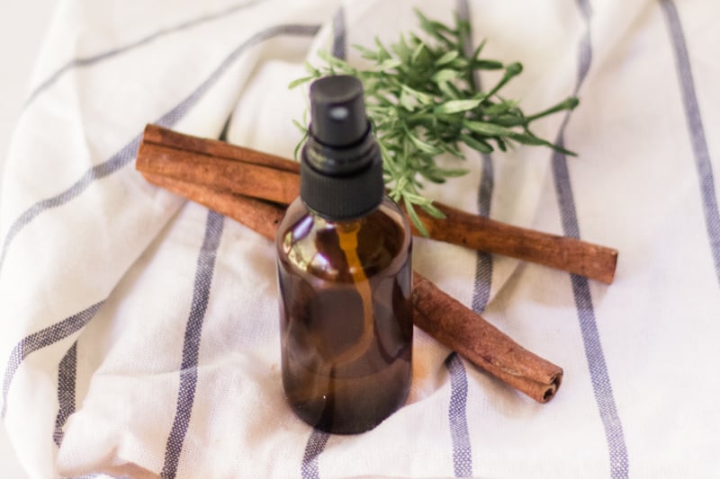 Homemade mouth freshener with essential oils in amber colored bottle with cinnamon sticks and rosemary sprigs behind it.