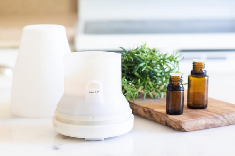 White essential oil diffuser on table with amber colored essential oil bottles and rosemary sprigs. 