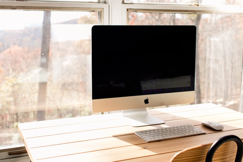 Apple desktop computer sitting on wood desk in front of window with a freshly cleaned screen.
