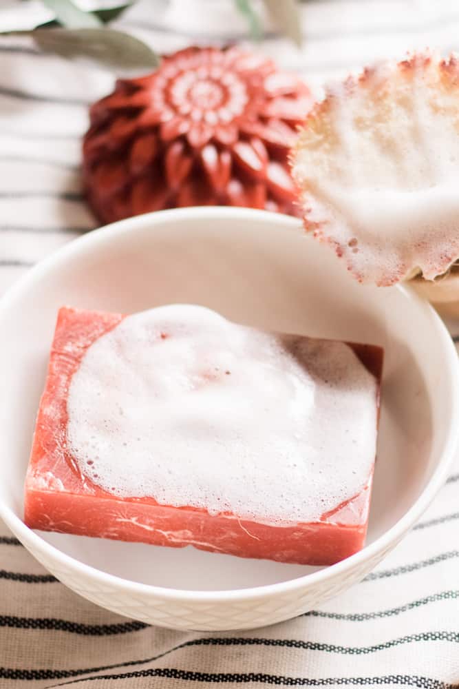 Red shave soap bar lathered up in a white bowl with a scrub brush next to it. 