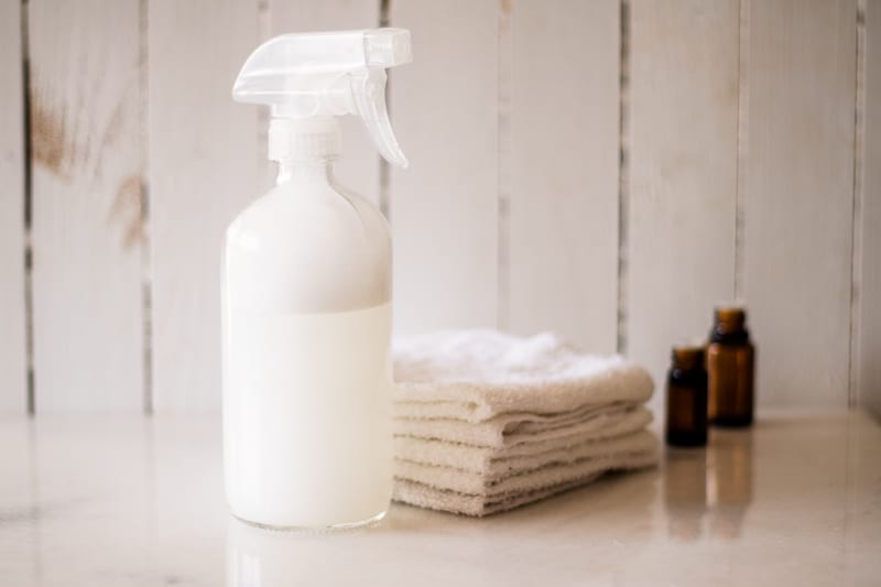 Glass spray bottle, white washcloths, and two amber colored essential oil bottles on white marble.