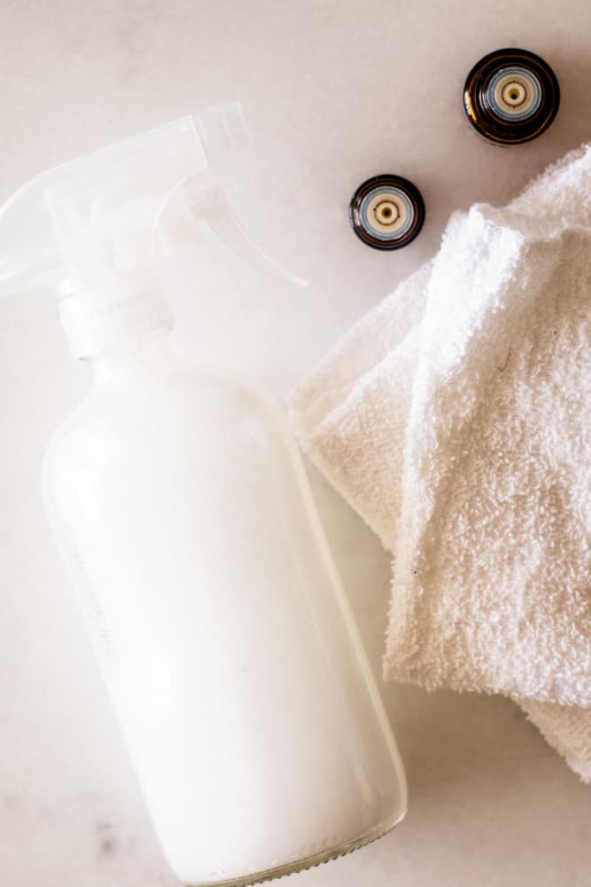 Glass spray bottle with cleaning solution and white cloths next to it.