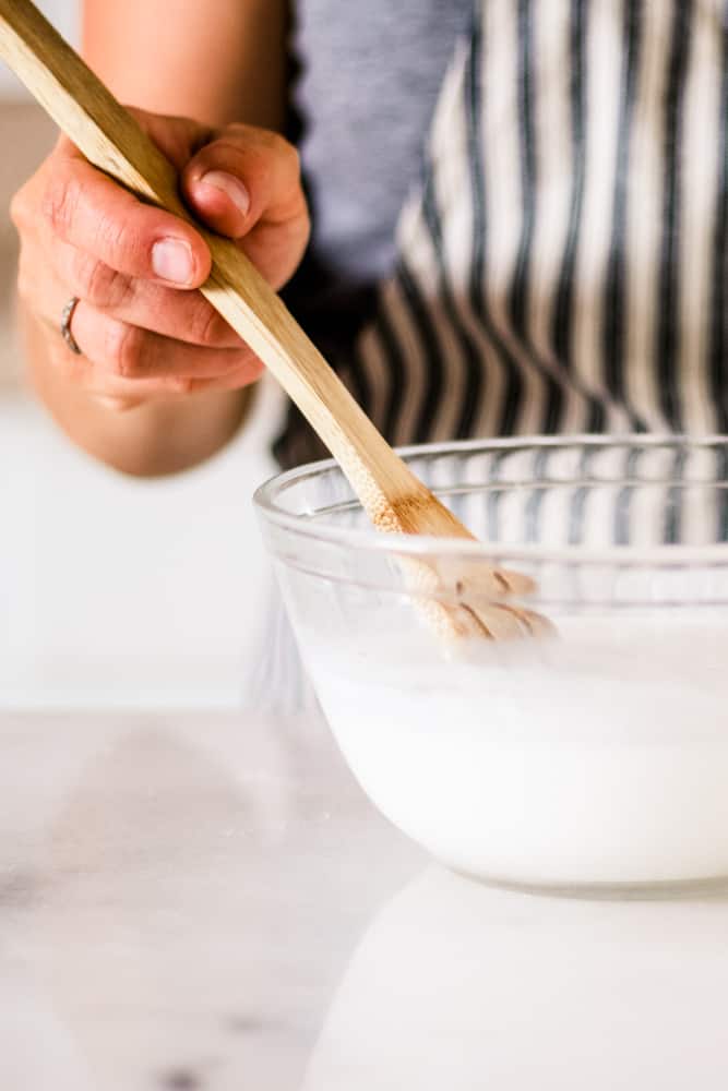 Stirring soap base after its melted in a clear glass bowl with a wooden spoon.