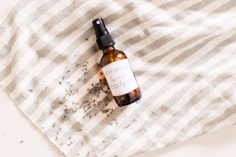 Homemade body oil in amber colored spray bottle laying on white and gray striped towel with lavender buds sprinkled around it.