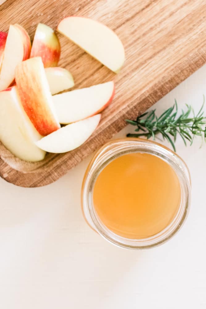 Apple cider vinegar shampoo, red apple slices, and sprigs of rosemary on white table. 