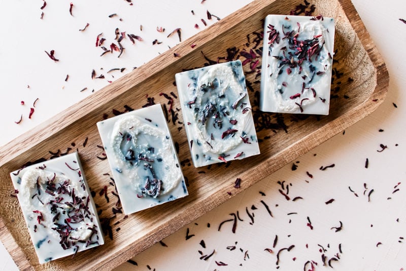 Loofah soap bars on a wooden soap tray garnished with hibiscus flowers.