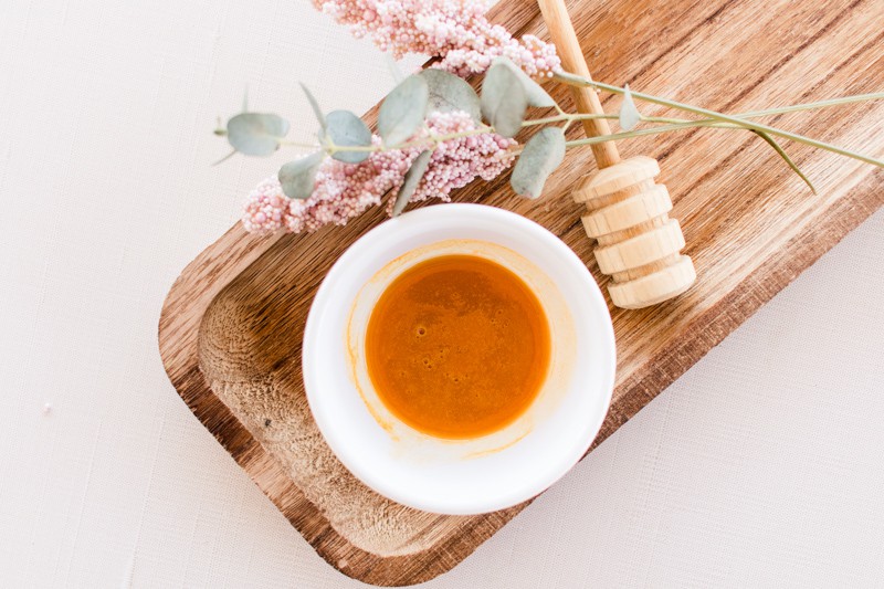 Glowing skin face mask with honey in a small white container, applicator and dried springs on white vanity.