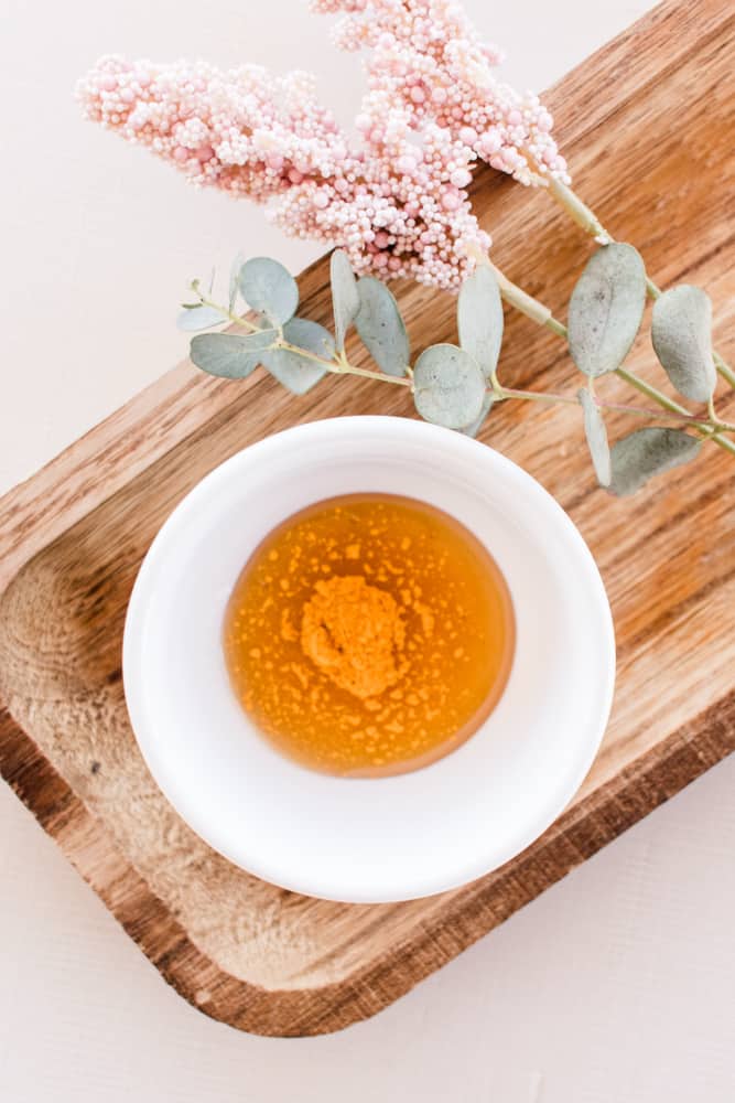 Homemade honey face mask in small white bowl with turmeric plant on wooden board.