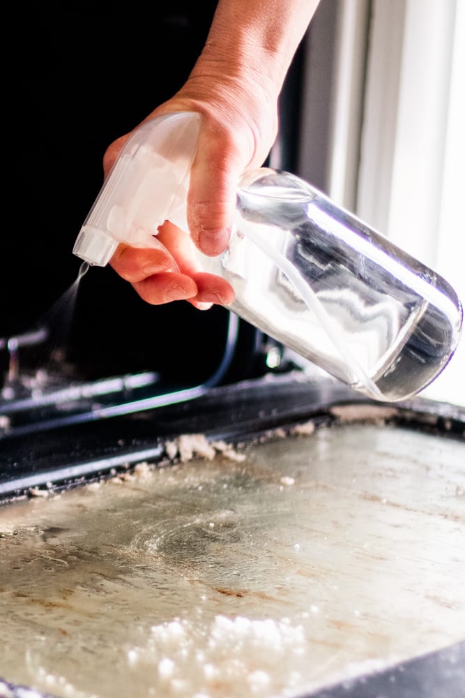 Homemade oven cleaner being sprayed onto oven door glass.