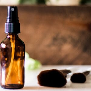 DIY makeup setting spray in glass spray bottle with applicator brushes on white marble vanity.
