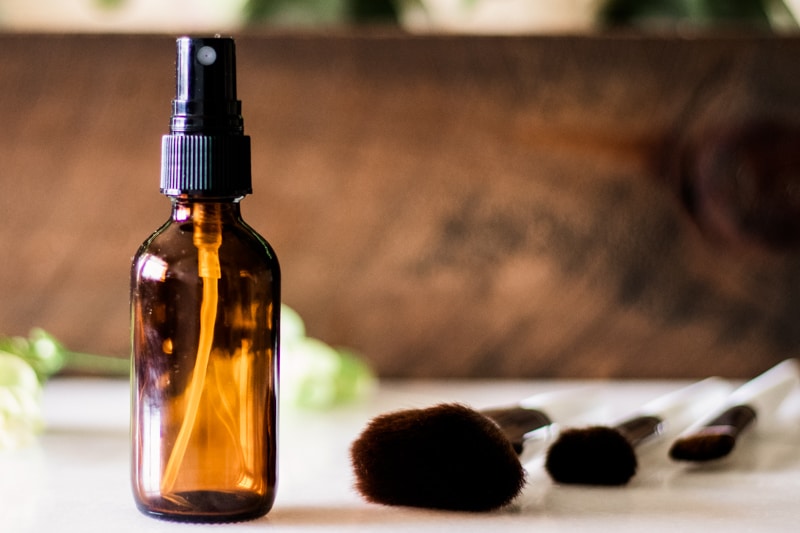 DIY makeup setting spray in glass spray bottle with applicator brushes on white marble vanity.