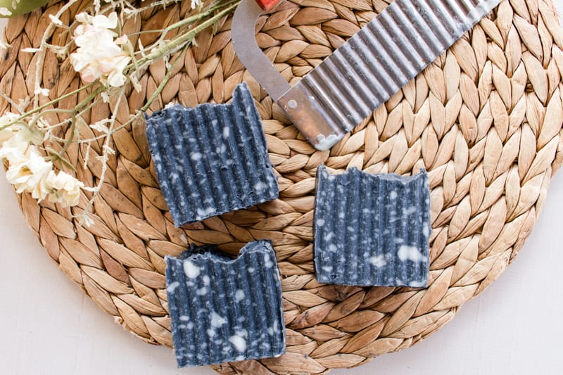 Activated charcoal soap bars with textured sides, a soap cutter, and lavender flowers on a wicker mat.