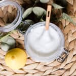 Soft scrub shower cleaner with a wooden spoon and a whole lemon on a wicker mat.