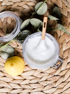 Soft scrub shower cleaner with a wooden spoon and a whole lemon on a wicker mat.