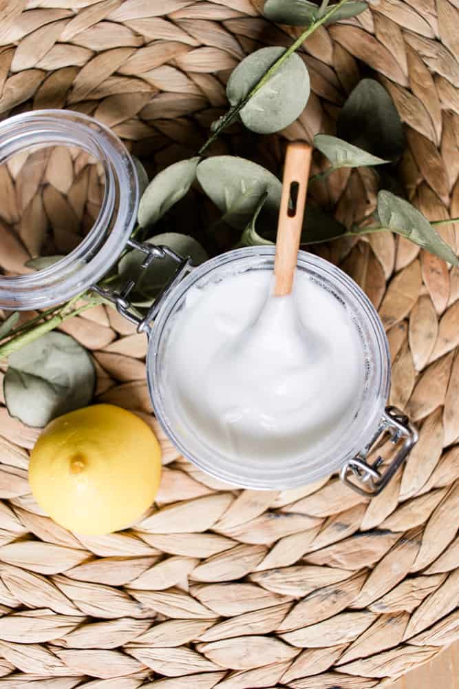 Soft scrub shower cleaner with a wooden spoon and a whole lemon on a wicker mat.