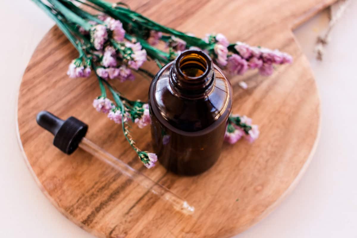 The mixed facial cleansing oil on a round wooden tray on a white vanity with colorful flowers next to it. 