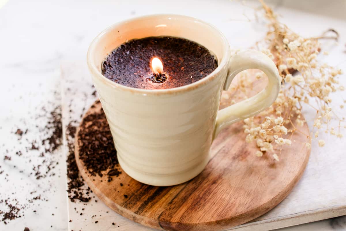 Homemade coffee candle burning in a pewter colored coffee mug with a decorative background.