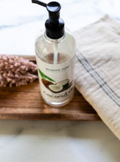 A bottle of fractionated coconut oil on a wooden tray on a white marble vanity.