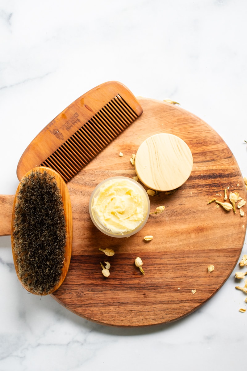 Beard butter, comb, brush all sitting on a wooden tray on a white vanity.