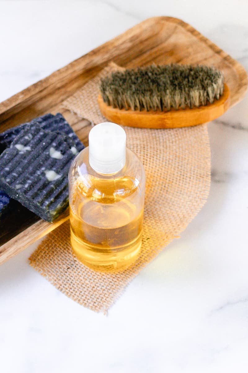 The DIY pre shave oil sitting in a bottle with a few bars of charcoal soap and a beard brush in the background.