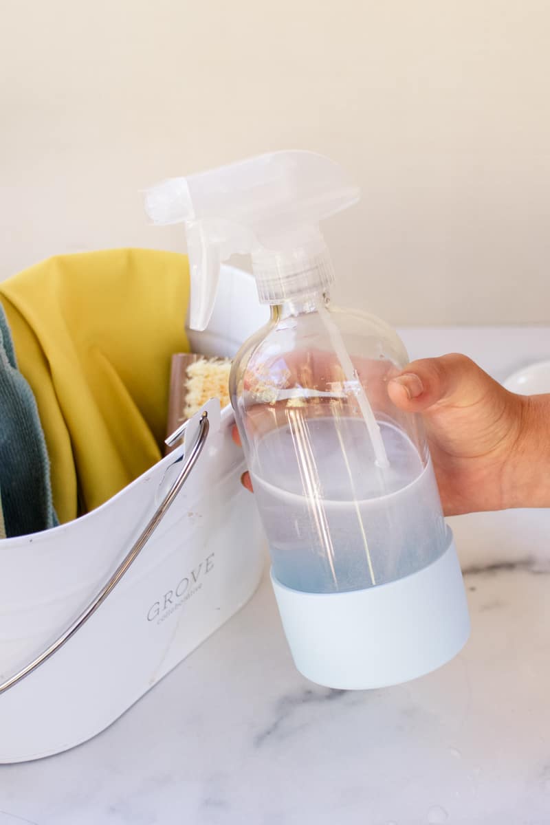 A bottle of homemade tile grout mold spray in front of a pail with rags and a brush.