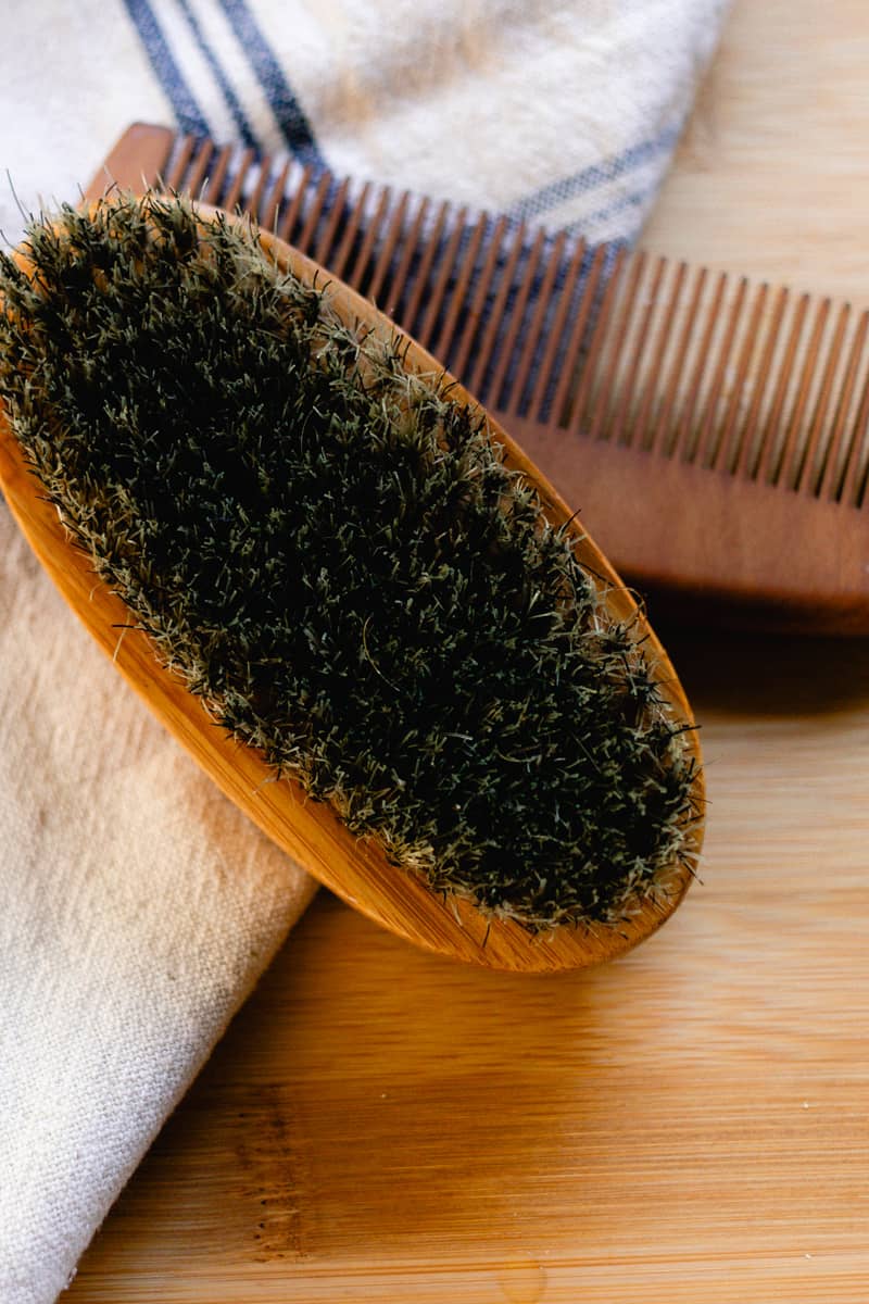 A clean boars bristle beard brush on a wooden top with a beard comb in the background.
