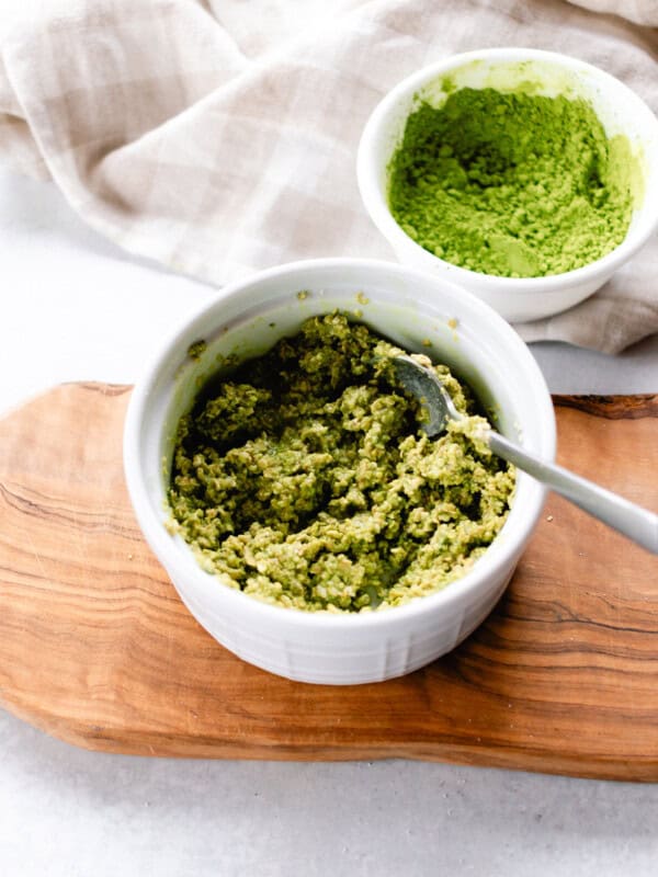 A green tea face mask in a white bowl being stirred with a metal spoon.