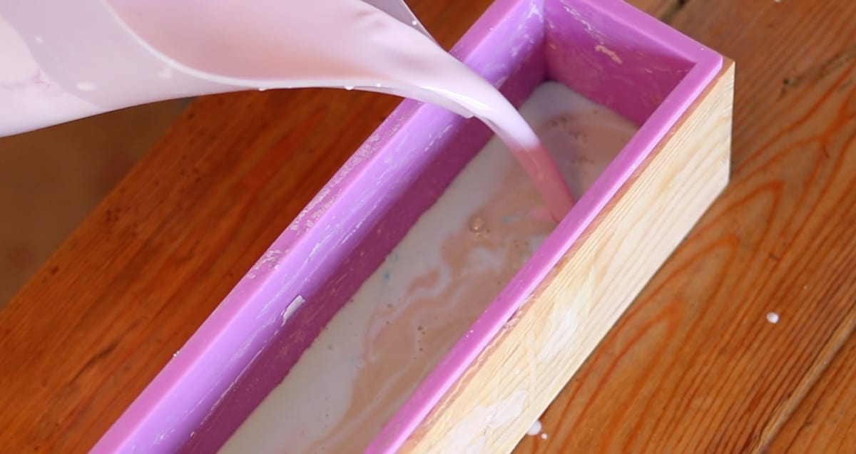 A plastic pitcher of the liquid mixture for DIY bar soap being poured into a silicone soap mold.