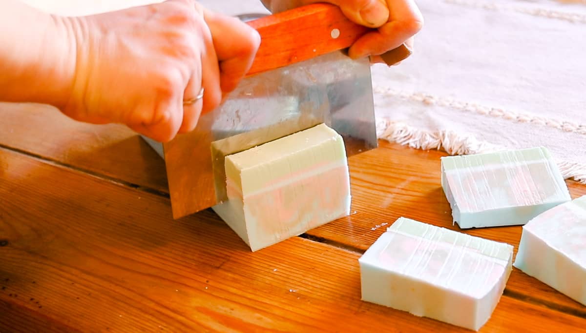 Two hands using a straight soap cutter to cut a loaf of colorful coconut oil bar soap into individual slices of bar soap.