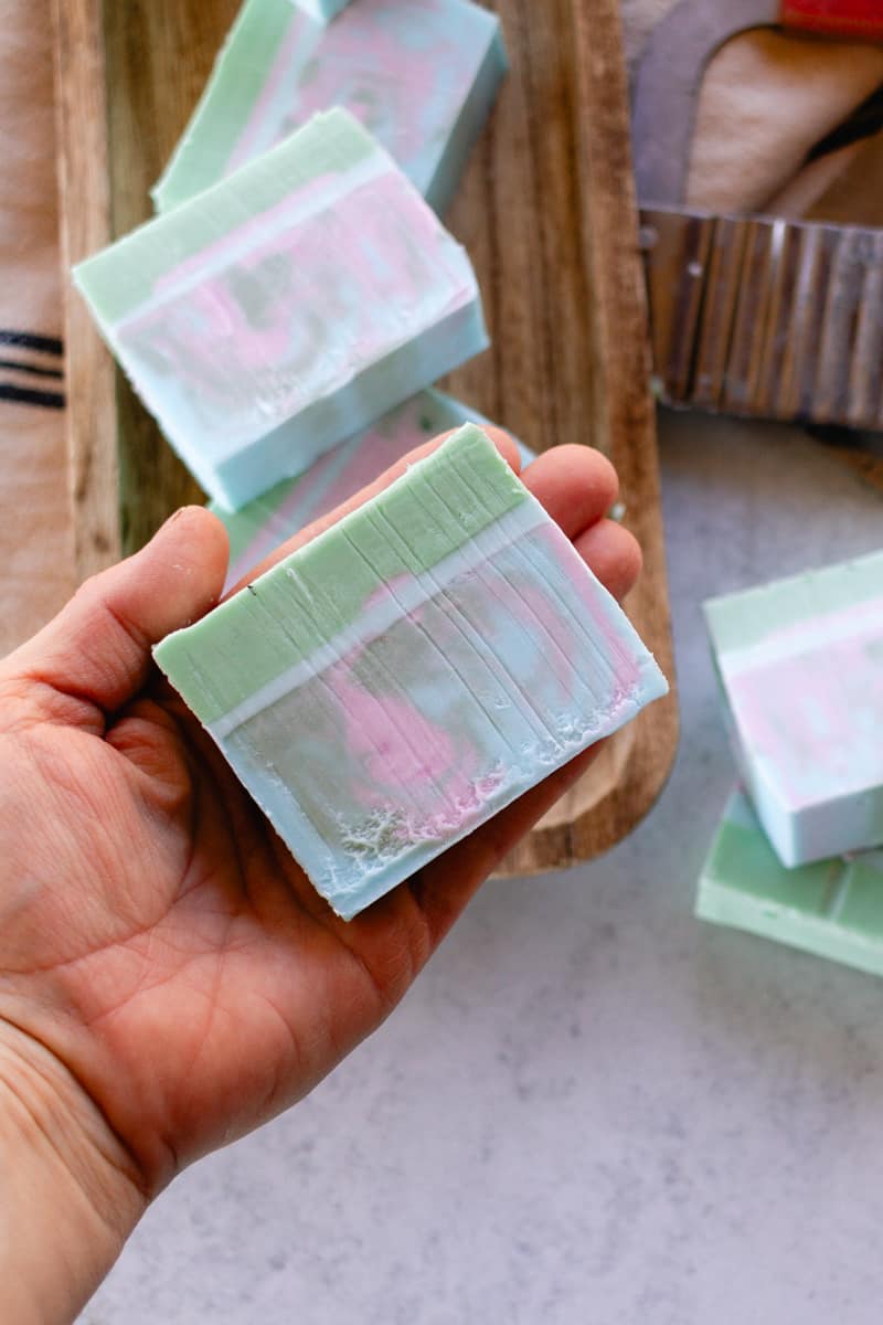 A hand holding a colorful homemade melt and pour coconut oil bar of soap with several bars in the background and a wavy soap cutter.